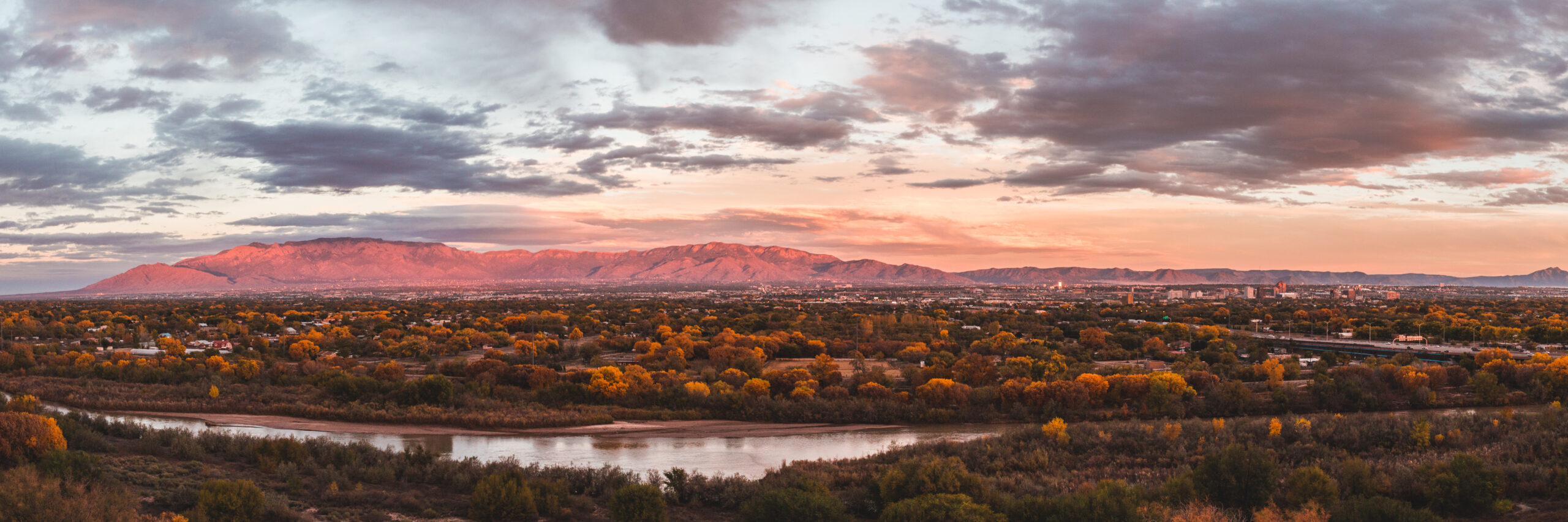 Panoramic view of Albuquerque in autumn with golden trees, the Rio Grande river in the foreground, and the Sandia Mountains glowing pink under a dramatic sunset sky.