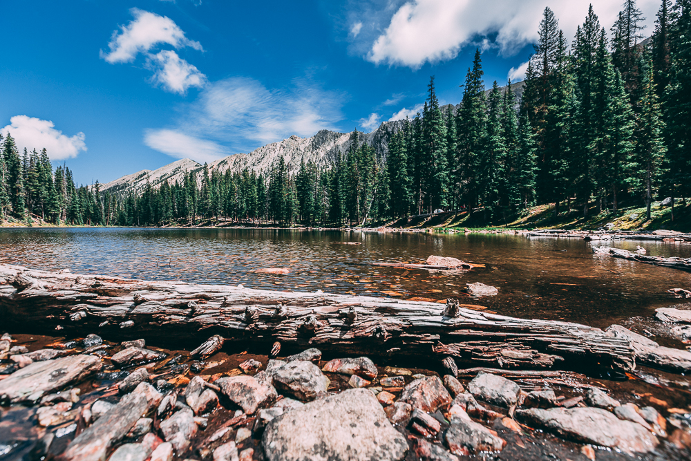 Time-lapse video of a mountain lake surrounded by forest and rocky terrain, showing clouds drifting rightward and water alternating between calm and wind-stirred ripples.