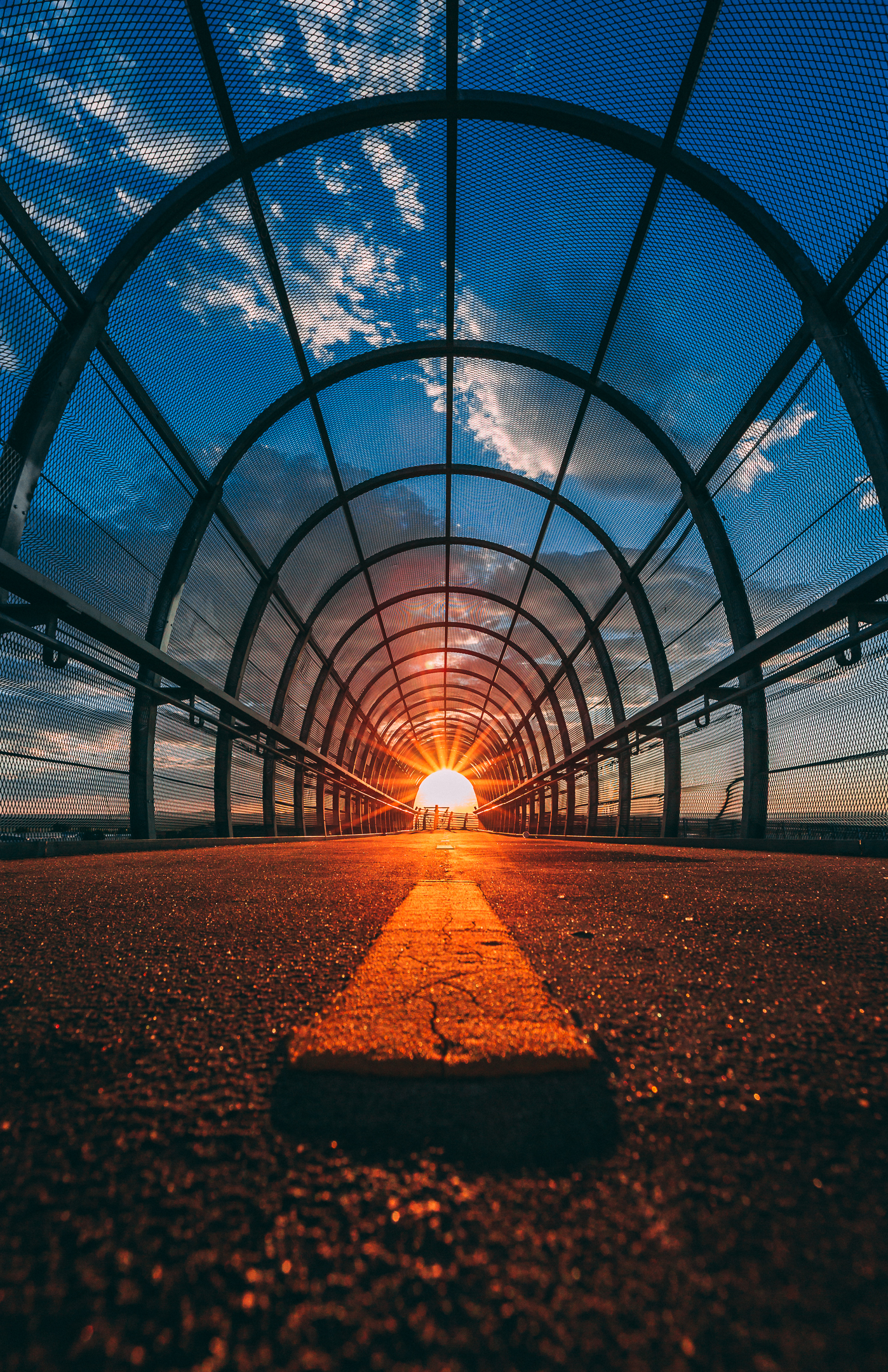 Symmetrical pedestrian tunnel with metal arches leading to a glowing sunset; warm tones and a central yellow line create depth and visual focus.