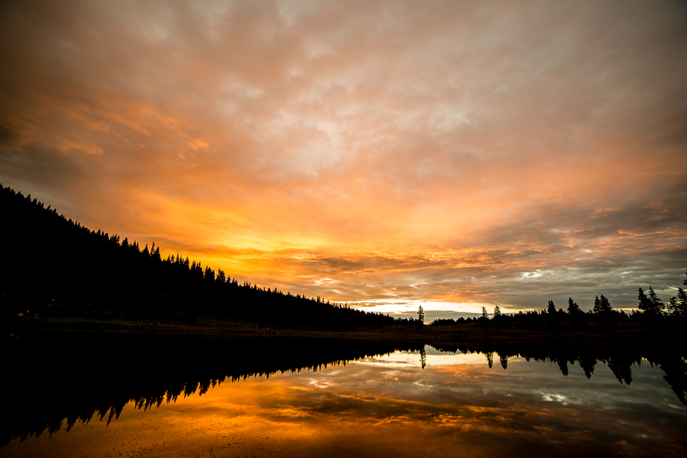 Timelapse of sunrise at Pecos Baldy Lake, showing vibrant sky colors reflected on calm water, with clouds moving in opposite directions and bug ripples on the lake surface.
